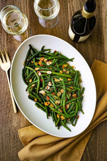 An overhead view of a large oval white platter holding green beans almondine with a bottle of wine and two glass of white wine nearby.