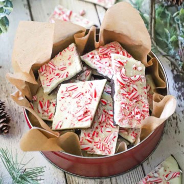A circular tin lined with brown paper filled with homemade peppermint bark, surrounded by holiday garland and more pieces of bark.