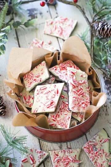 A circular tin lined with brown paper filled with homemade peppermint bark, surrounded by holiday garland and more pieces of bark.