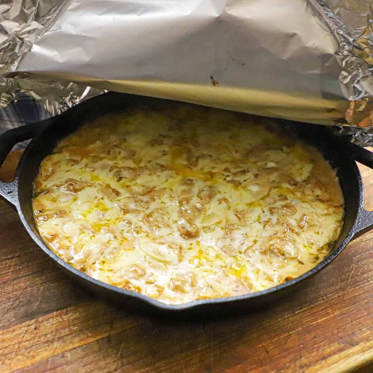 A person removing a layer of aluminum foil from the top of a large black cast-iron skillet that is filled with a partially baked French onion au gratin potatoes.