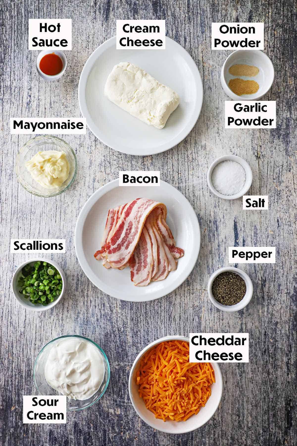 An arrangement of ingredients for Ultimate Hot Bacon and Cheddar Dip on a grey wooden background including strips of uncooked bacon, cream cheese, mayonnaise, cheddar cheese, sour cream, scallions, and seasonings.