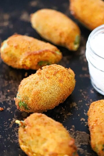 Several freshly fried jalapeño poppers on a black baking sheet next to a small glass jar filled sour cream.