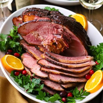 A large white platter filled with a honey mustard glazed smoked ham that has been partially sliced and with fresh herbs and orange slices garnishing the sides of the dish.