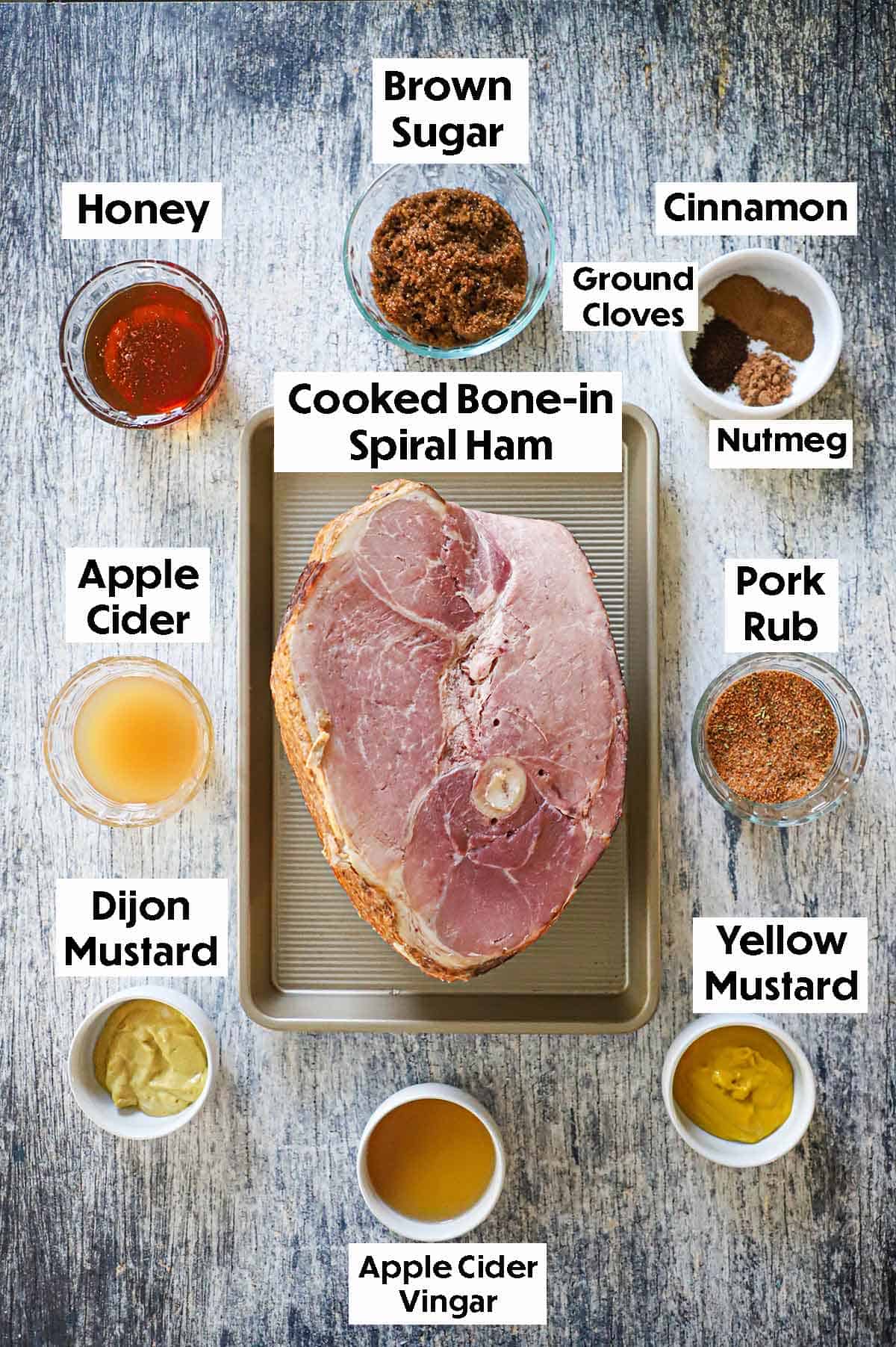 An arrangement of ingredients for a honey mustard glazed smoked ham on a grey wooden background including a cooked spiral ham, honey, brown sugar, apple cider, spices, Dijon mustard, yellow mustard, and apple cider vinegar.