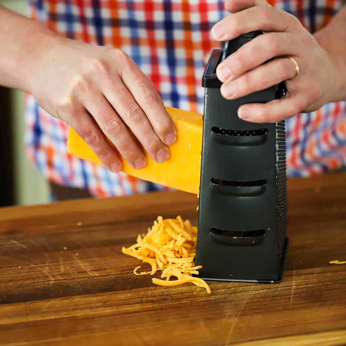 A person grating a block of cheddar cheese on a box grater on a wooden cutting board.