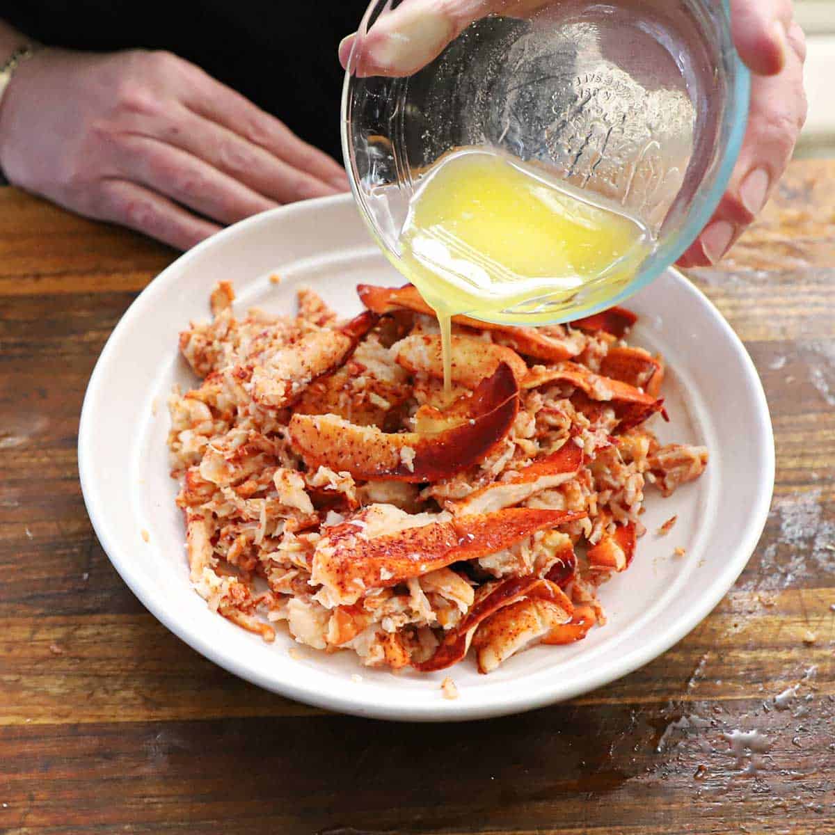A person pouring melted butter from a small glass bowl over chopped cooked lobster meat in a white bowl on a cutting board.