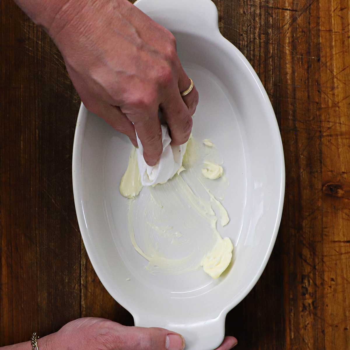 A person using paper towels to spread softened butter all over the inside of a white oval baking dish.