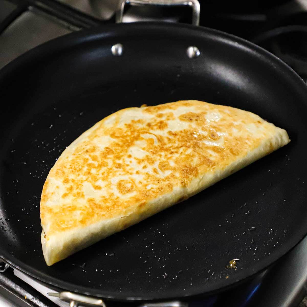 A large steak fajita quesadilla that is being lightly browned in a large non-stick skillet with a little oil over a gas flame.
