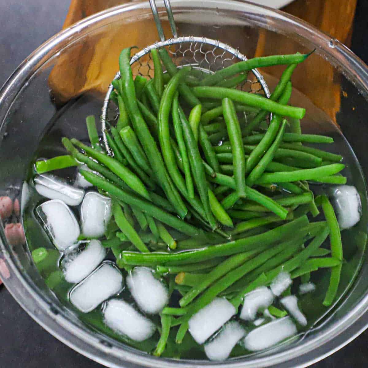 A person holding a large glass bowl filled with simmered fresh green beans in an ice bath.