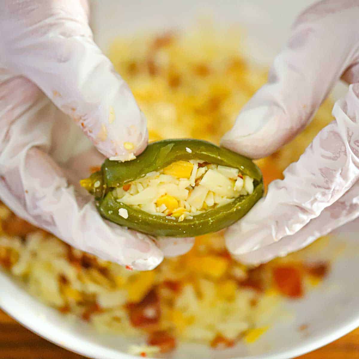 A person holding a whole pickled jalapeño that has been split down the side and has been stuffed with shredded cheese and crispy bacon pieces.