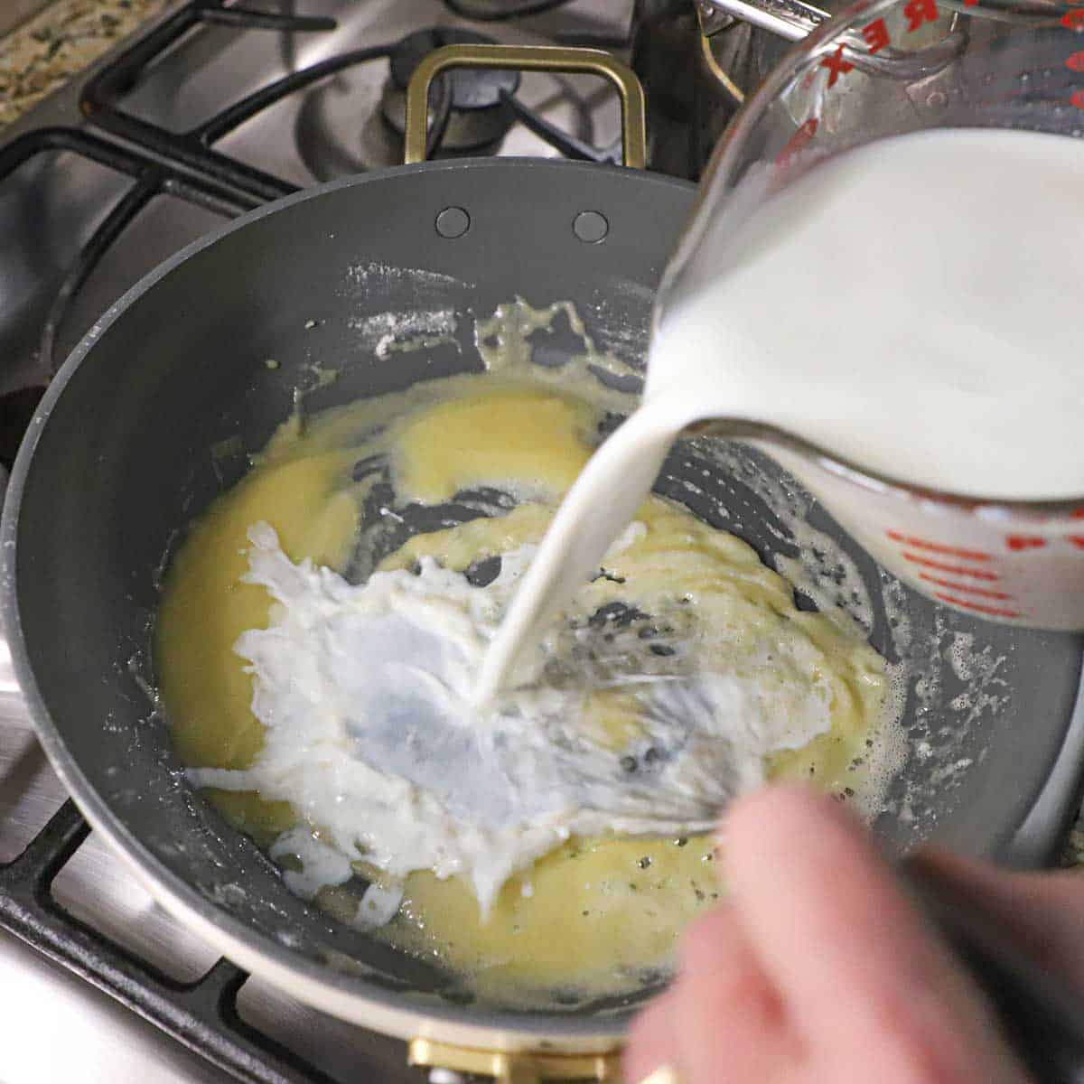 A person whisking whole milk that is poured into a skillet filled with an amber roux on a gas stove.
