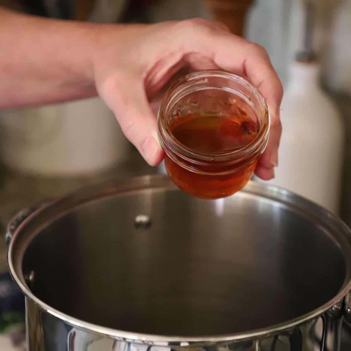 A person holding a small glass jar filled with maple syrup of a silver stock pot filled with mulled wine on a gas stove.