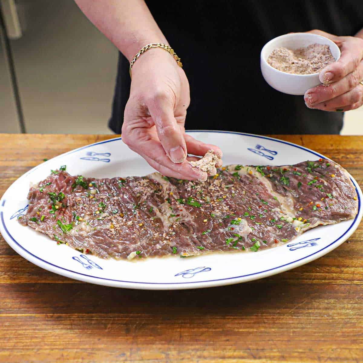 A person sprinkling fajita seasoning over the surface of an uncooked marinated skirt steak that is resting on a large white platter on a cutting board.