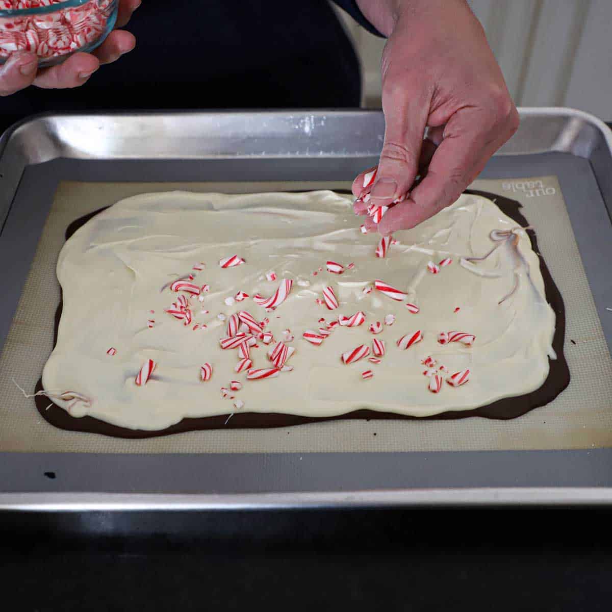 A person sprinkling crushed candy canes over the surface of a rectangular shape of peppermint bark before it has been chilled.