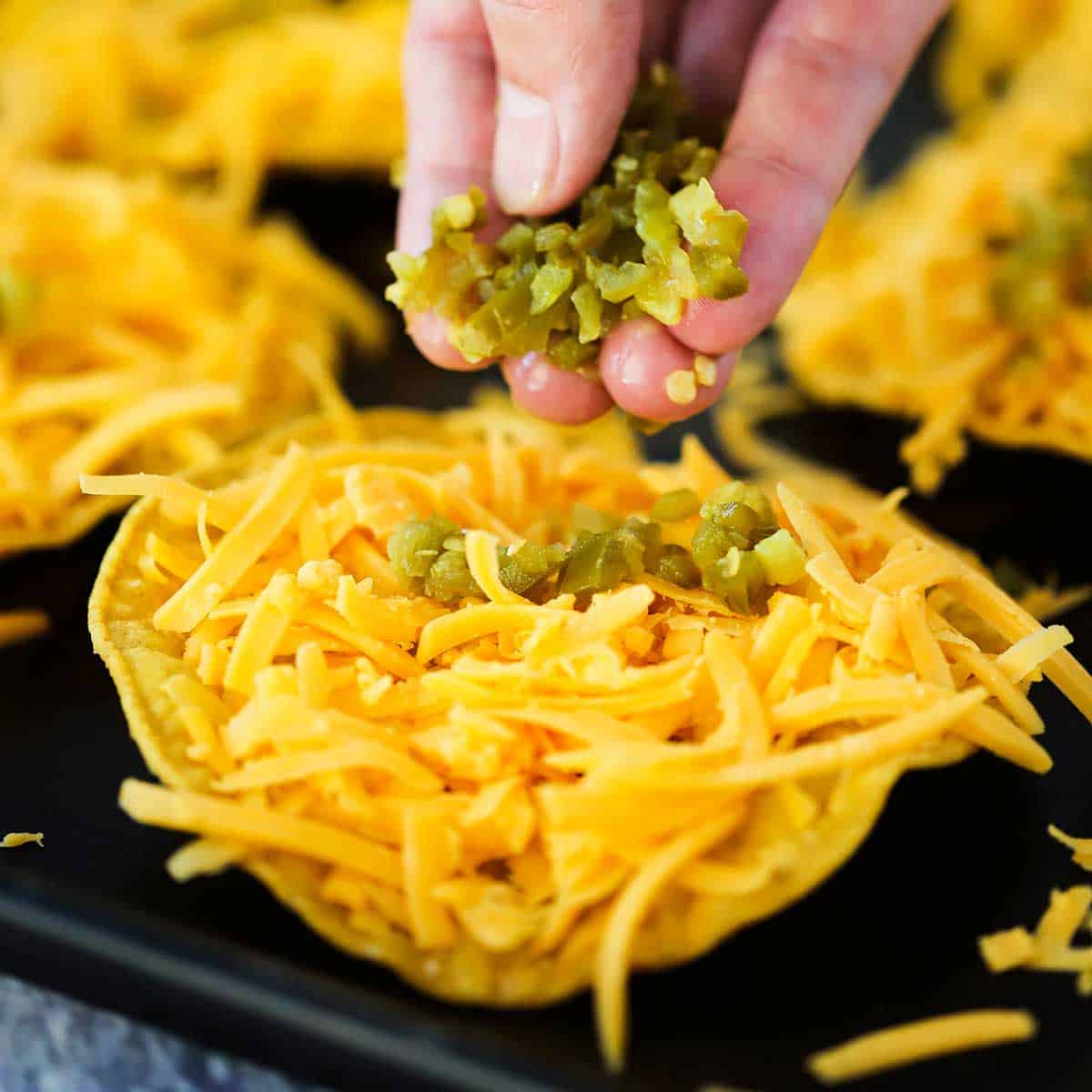 A person sprinkling chopped jalapeños over the top of a corn tortilla that is covered with shredded cheddar cheese.