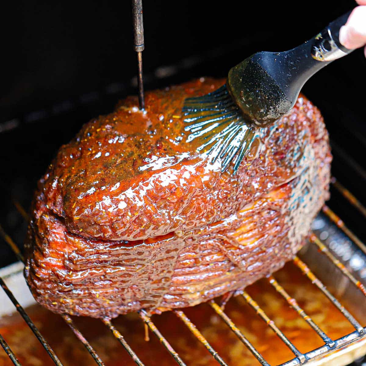 A person using a large grill brush to apply a thick layer of glaze over a smoked spiral-sliced ham being smoked inside an electric smoker.