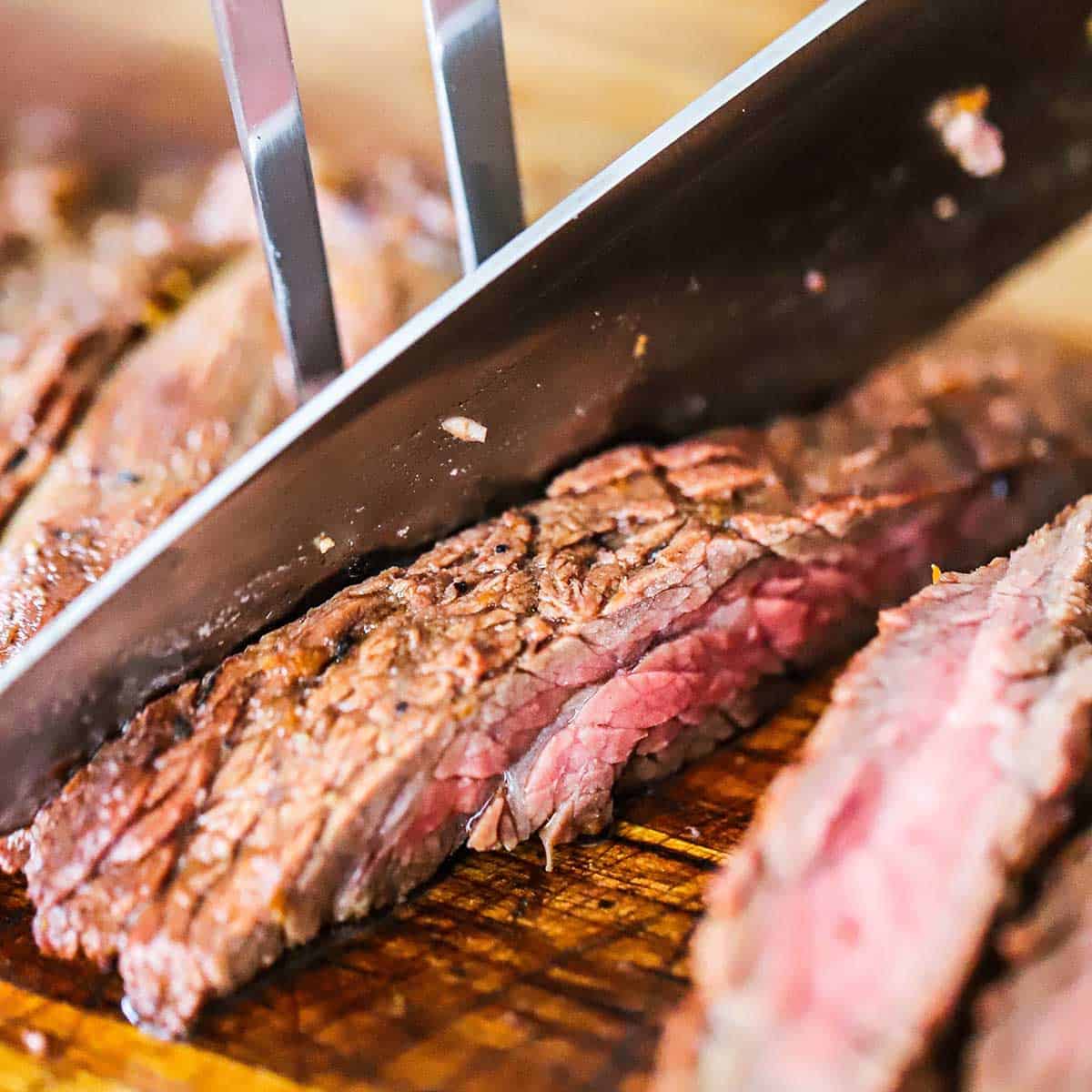 A close-up view of a person using a large fork and chef's knife to slice freshly grilled fajita steak on a wooden cutting board.