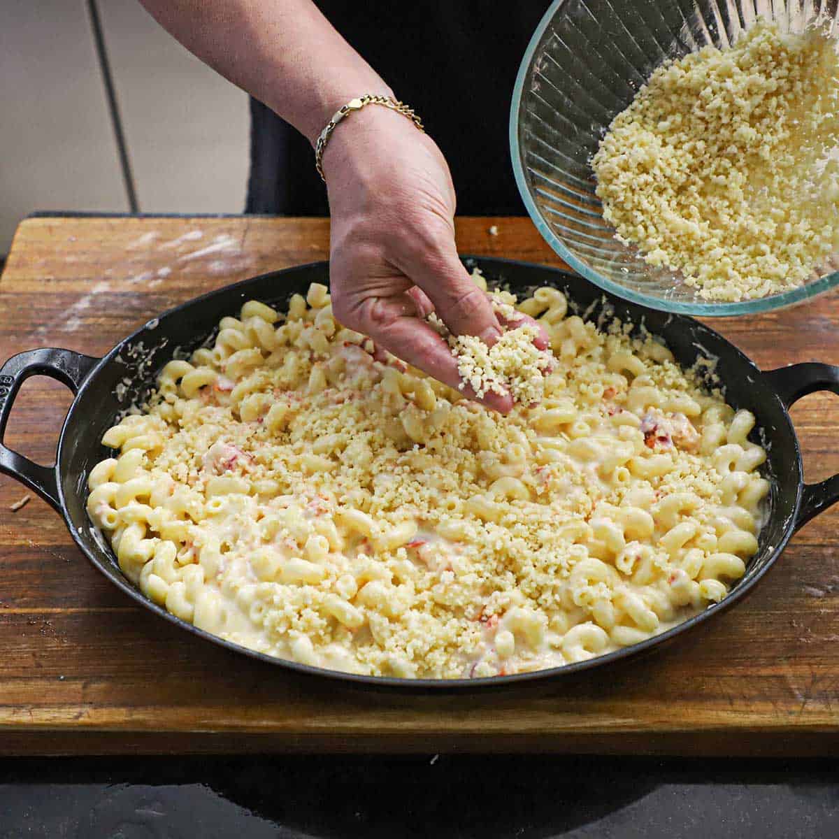 A person using his hand to sprinkle a buttery Panko and Parmesan mixture over the top of a dish filled with homemade lobster mac and cheese that has not been baked yet.