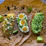 Three avocado toasts sitting next to each other on a crumpled brown paper, with one being a classic avocado toast, and the one in the middle topped with sliced soft-boilded eggs and herbs, and the last one being a Mexican street corn, eloté, version.