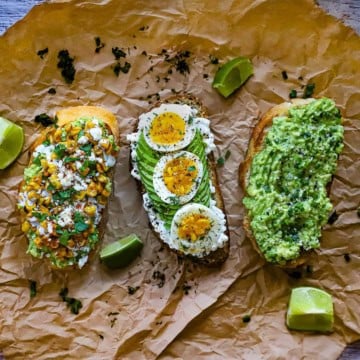 Three avocado toasts sitting next to each other on a crumpled brown paper, with one being a classic avocado toast, and the one in the middle topped with sliced soft-boilded eggs and herbs, and the last one being a Mexican street corn, eloté, version.