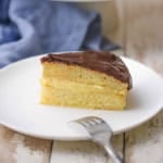 A close-up view of a slice of Classic Boston Cream Pie sitting on a white dessert plate with a fork resting nearby.