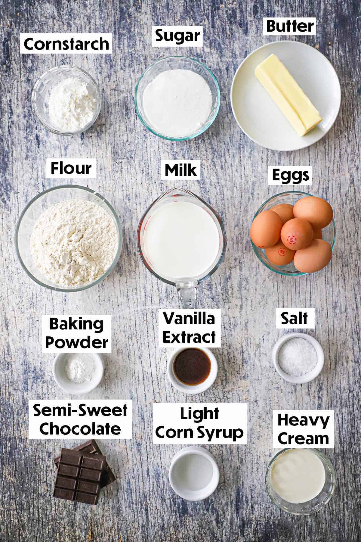 An arrangement of ingredients for Classic Boston Cream Pie on a grey wooden background including flour, milk, eggs, butter, sugar, cornstarch, baking powder, vanilla extract, salt, chocolate, cory syrup, and heavy cream.