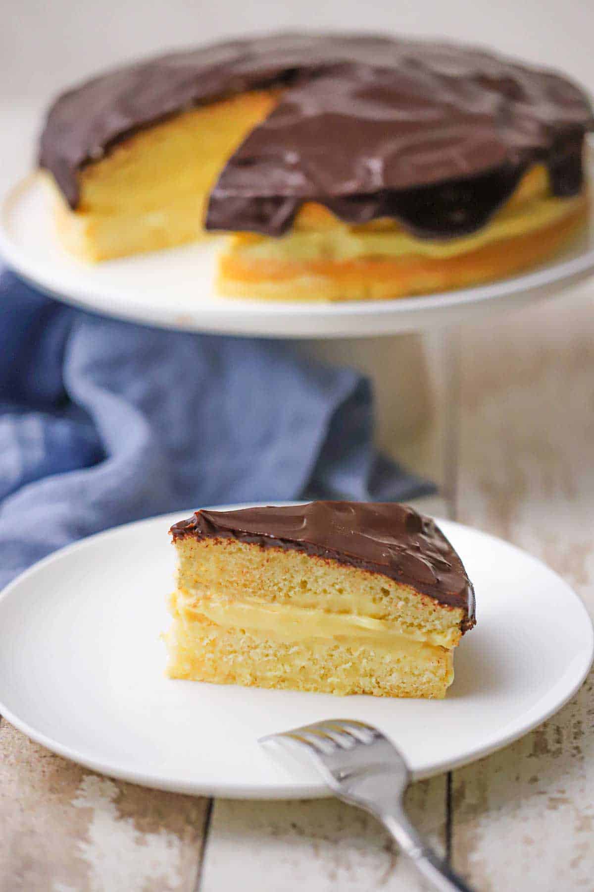 A slice of classic Boston cream pie resting on a white dessert plate with the entire dessert on a cake stand in the background.
