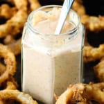 A close-up view of small glass jar filled with Cajun remoulade with a spoon in the middle of it and the jar is in the middle of a pile of homemade onion rings.