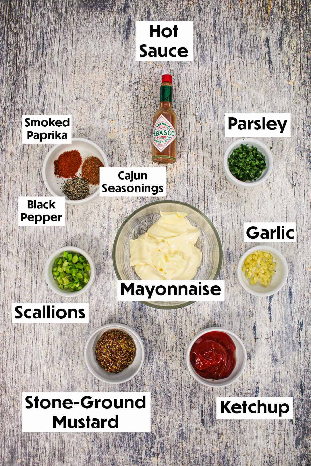 An arrangement of ingredients for Cajun remoulade on a grey wooden background including mayonnaise, ketchup, mustard, garlic, scallions, parsley, and spices.