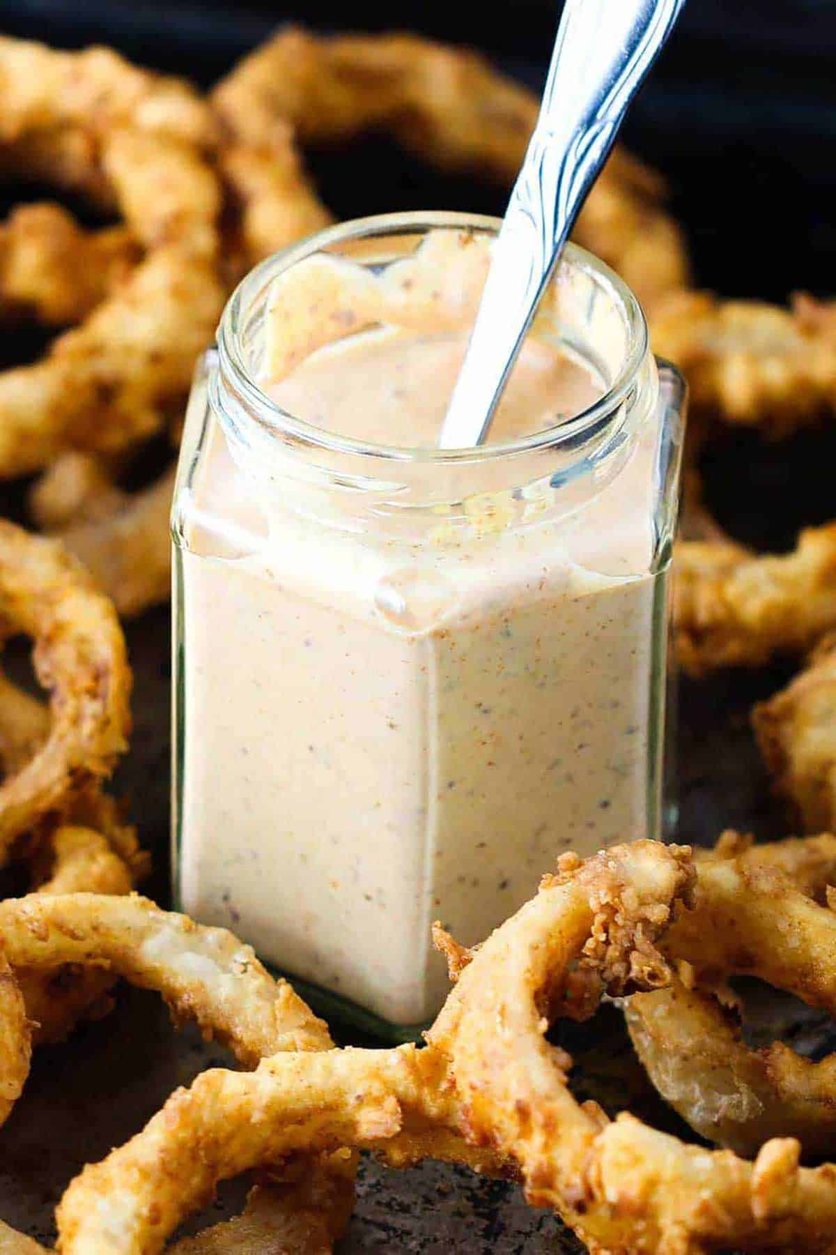 A close-up view of small glass jar filled with Cajun remoulade with a spoon in the middle of it and the jar is in the middle of a pile of homemade onion rings.