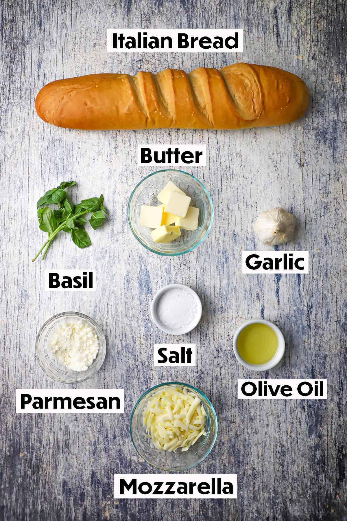 An arrangement of ingredients for cheesy garlic bread on a wooden grey background including a loaf of Italian bread, butter, basil, garlic, Parmesan cheese, olive oil, mozzarella, and salt.