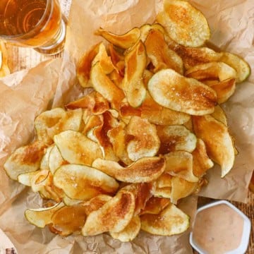 An overhead view of a pile of crispy homemade potato chips resting on crumpled brown paper next to a glass of beer and a small bowl filled with a dipping sauce.