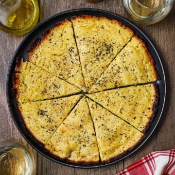 An overhead view of easy homemade socca that has been cut into triangles sitting in a large black plate next to two glasses of white wine.