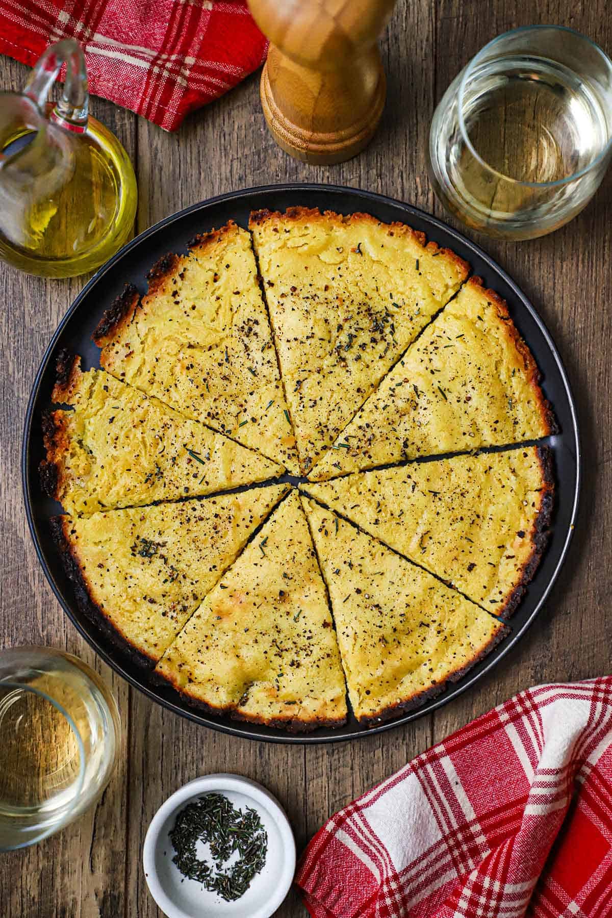 An overhead view of easy homemade socca that has been cut into triangles sitting in a large black plate next to two glasses of white wine and a red checkered napkin.