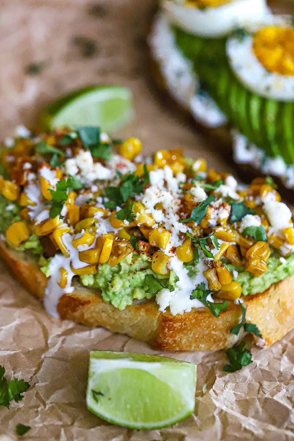 A close-up view of an Eloté avocado toast with a slice of toasted sourdough bread on the bottom, then topped with smashed avocado, charred corn, cream, Cotija cheese, and chopped cilantro.