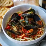 A white pasta bowl filled with a serving of Easy Mussels Marinara on top of cooked linguine with toasted bread and a glass of white wine nearby.