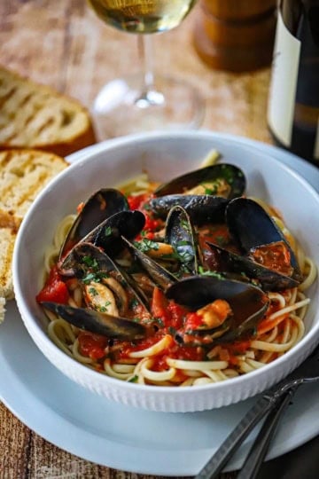A white pasta bowl filled with a serving of Easy Mussels Marinara on top of cooked linguine with toasted bread and a glass of white wine nearby.