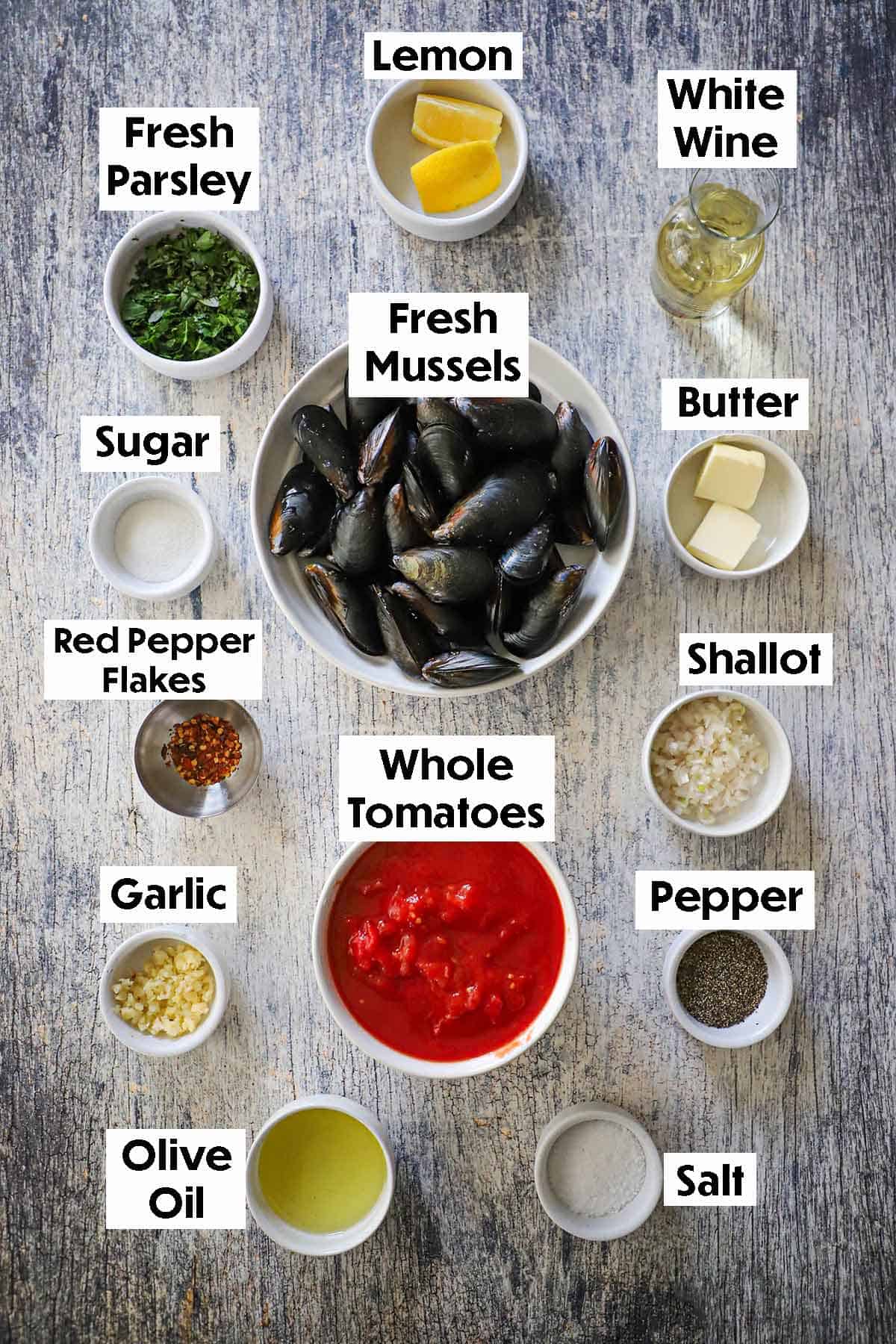 An arrangement of ingredients for Easy Mussels Marinara on a grey wooden background including fresh mussels, whole tomatoes in sauce, white wine, lemon, butter, shallots, garlic, herbs, and spices.
