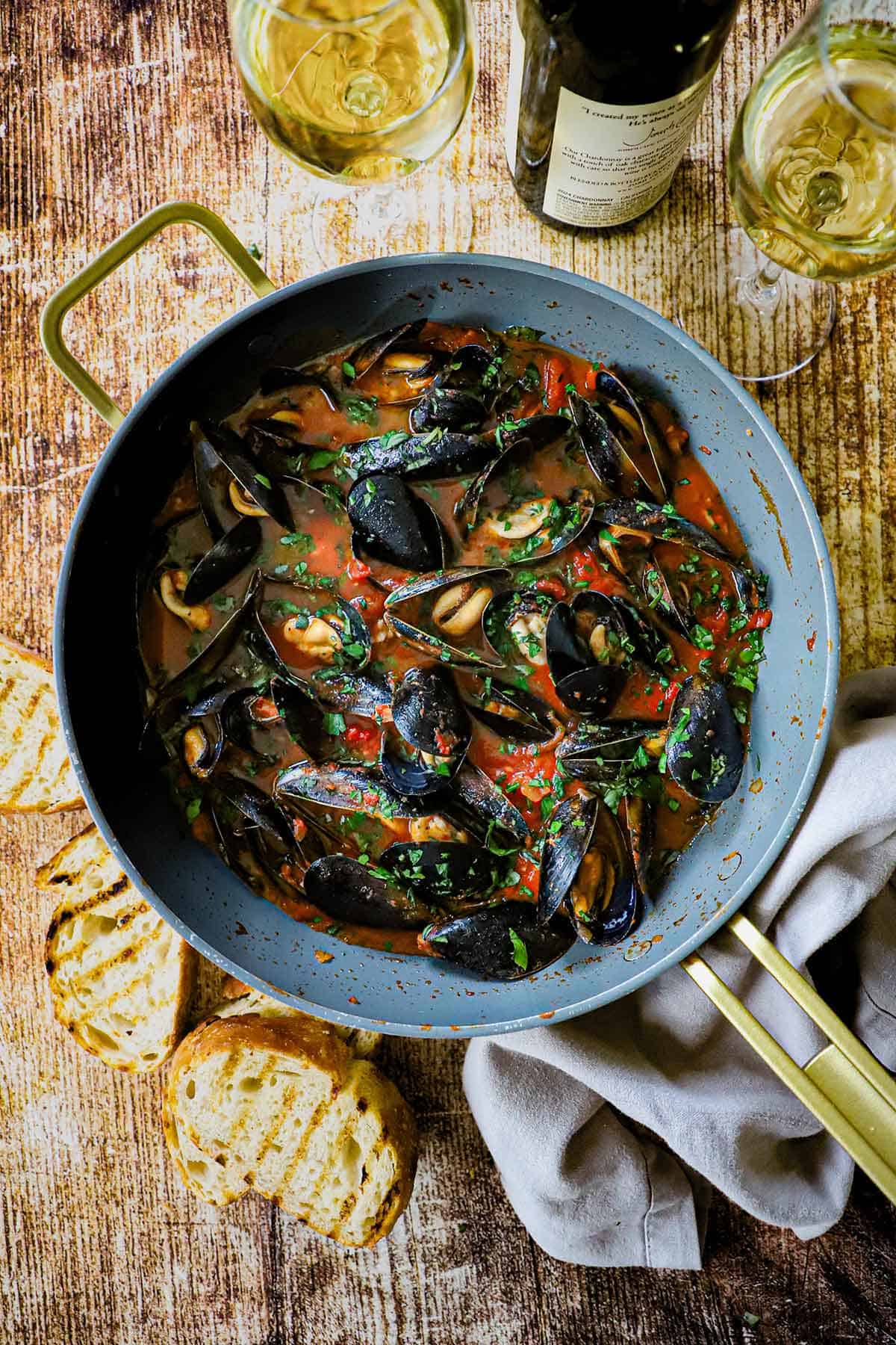 An overhead view of Easy Mussels Marinara in a large grey non-stick skillet with slices of toasted bread, two glasses of white wine, and a bottle of wine all nearby.
