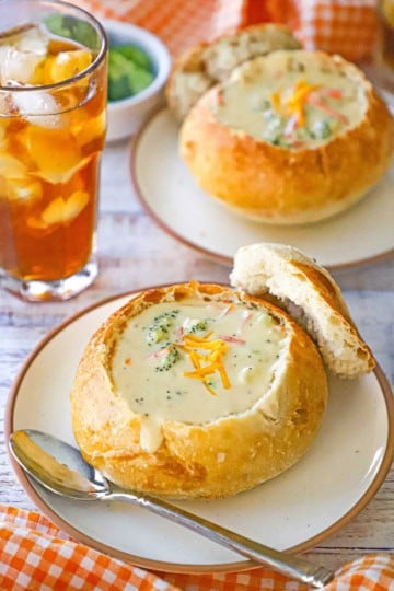Two sourdough bread bowls filled with Broccoli Cheddar Soup resting on small plates with a glass of iced tea nearby.
