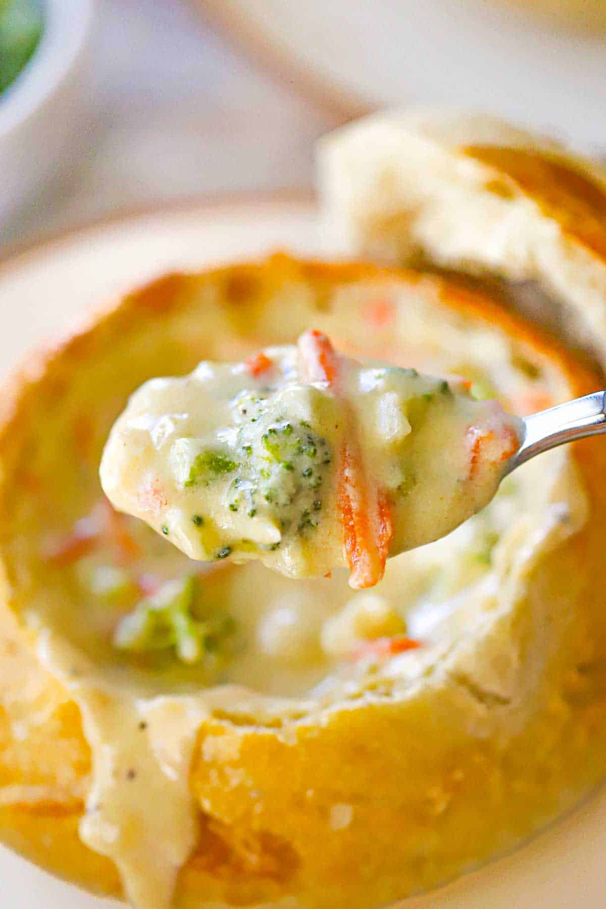 A spoon being used to raise a spoonful of broccoli cheddar soup over a bread bowl filled of the same.