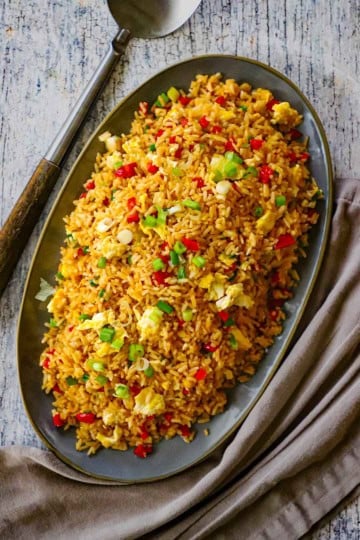 An overhead view of a grey oval platter holding a large mound of homemade Peruvian fried rice with a large serving spoon and grey napkin nearby.