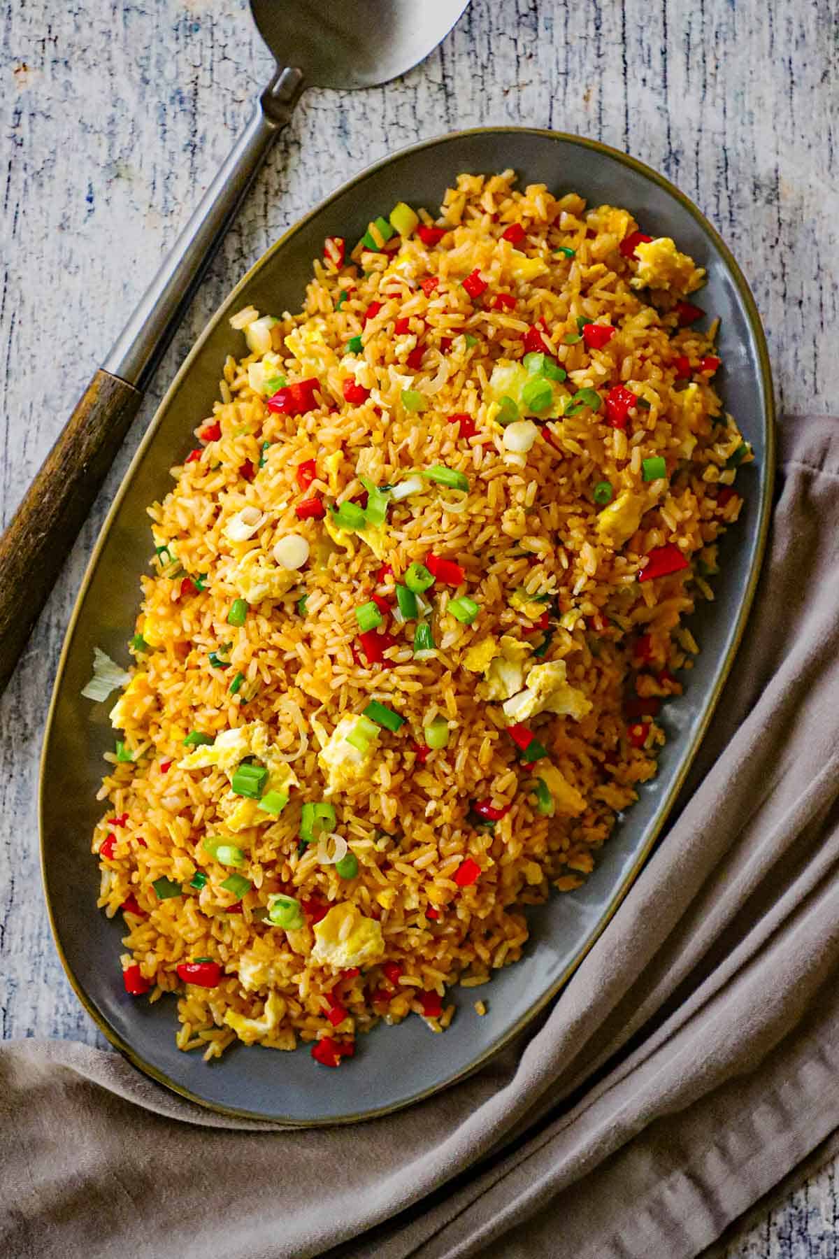 An overhead view of a grey oval platter holding a large mound of homemade Peruvian fried rice with a large serving spoon and grey napkin nearby.