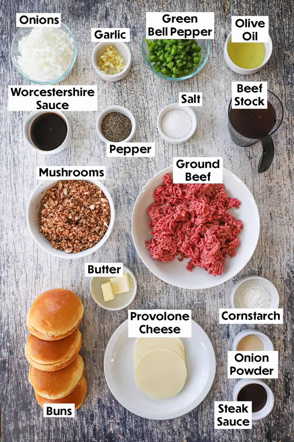 An arrangement of ingredients for Ultimate Philly Cheesesteak Sloppy Joes on a grey wooden background including ground beef, chopped mushrooms, onions, green bell pepper, garlic, beef stock, oil, butt, buns, provolone cheese, cornstarch, and seasonings.