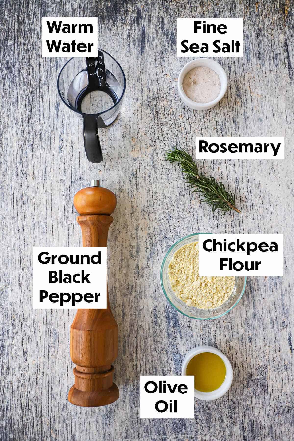 An arrangement of ingredients of ingredients for easy homemade socca on a wooden grey background including chickpea flour olive oil, water, sea salt, black pepper, and fresh rosemary.