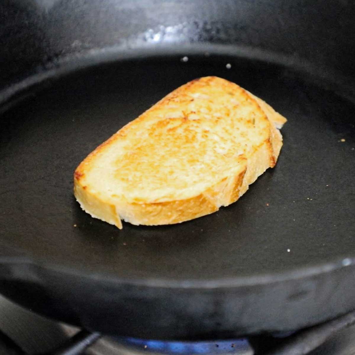 A slice of sourdough bread being toasted in a large cast-iron skillet over a gas stove.
