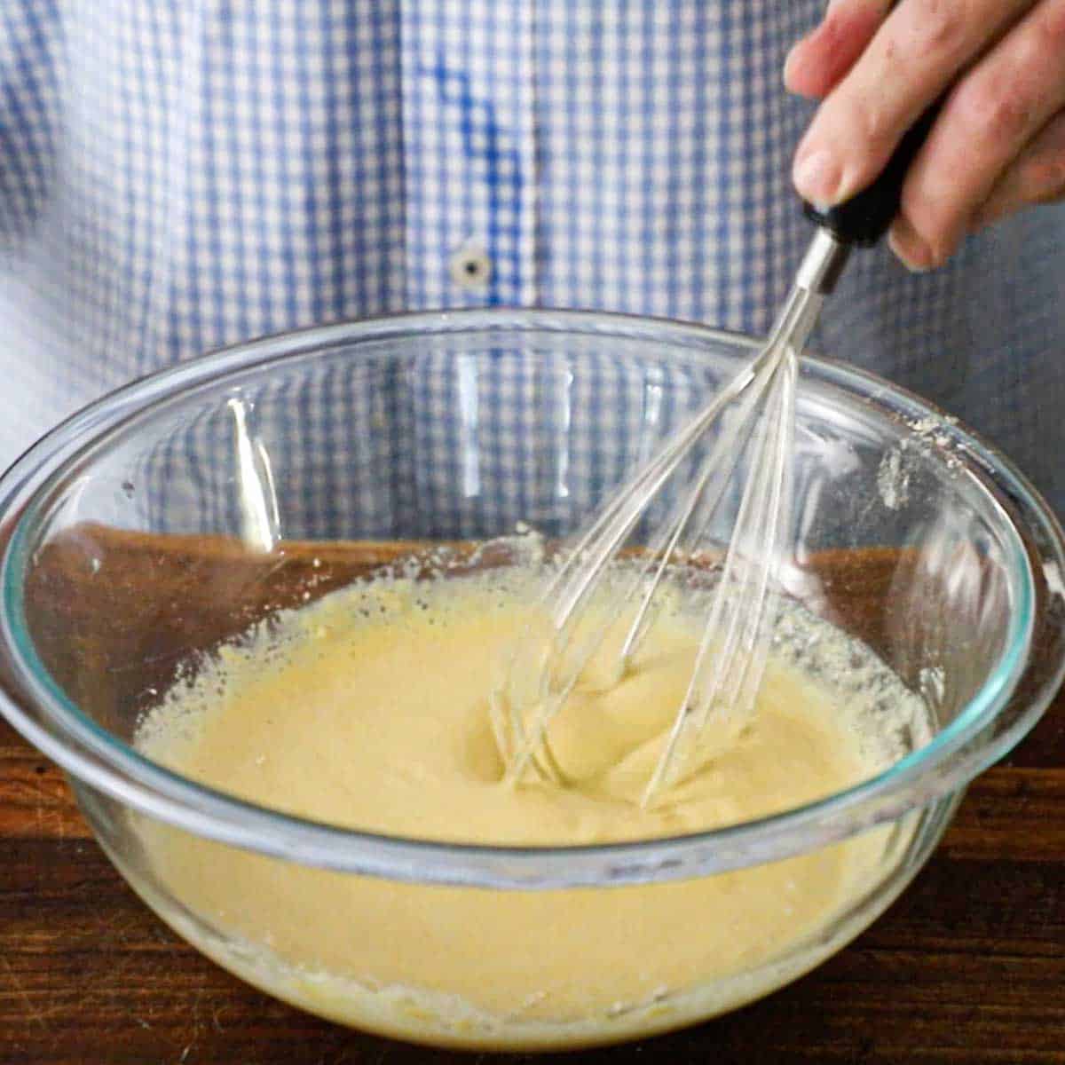 A person using a whisk to mix chickpea flour with water and olive oil in a large glass bowl.