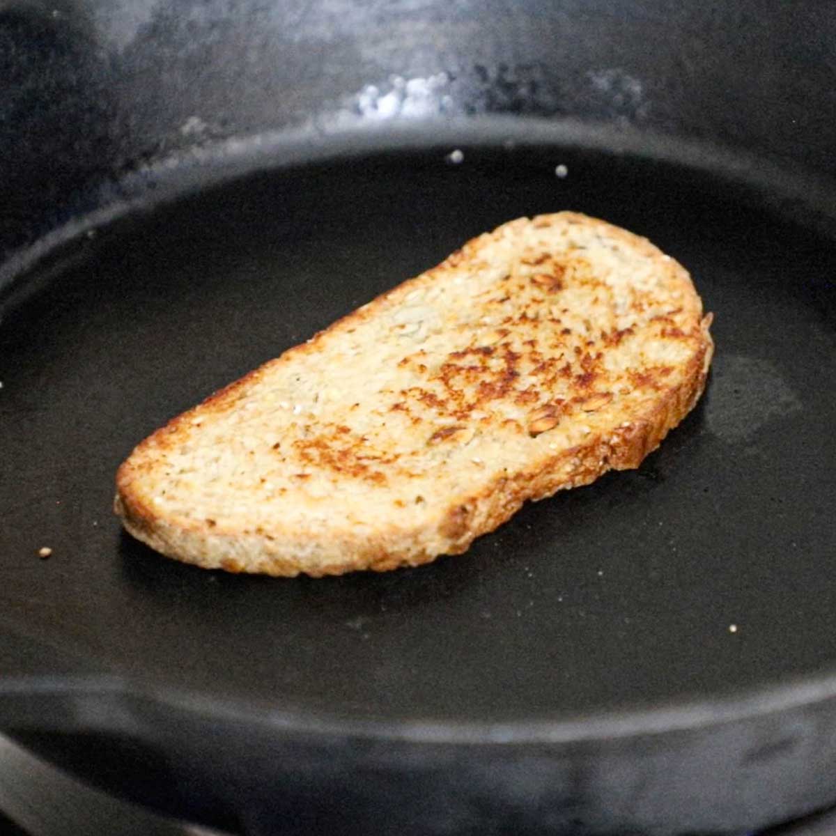 A slice of multi grain bread being toasted in a large cast-iron skillet on a gas stove.