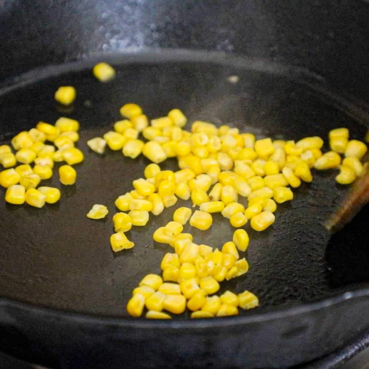 Sweet corn kernels being roasted in a hot cast-iron skillet on a gas stove.