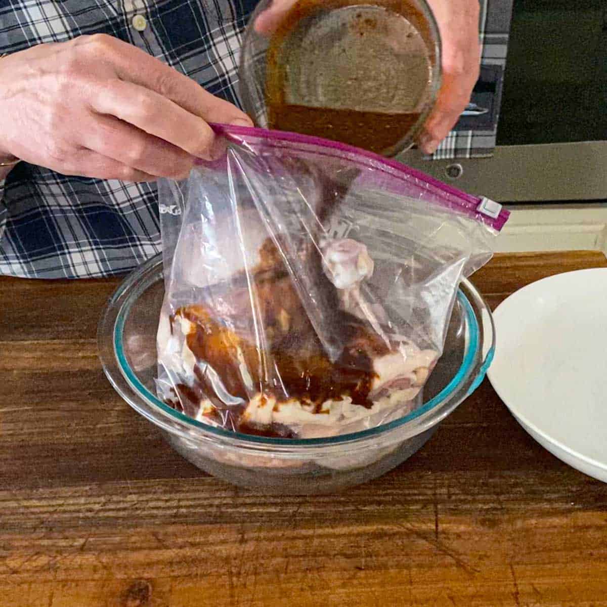 A person pouring a dark brown marinade into a large freezer baggie filled with chicken thighs and chicken legs.